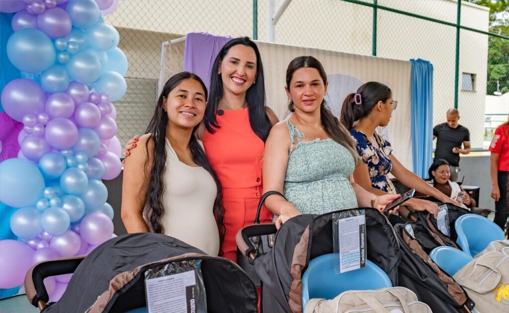 Três mulheres sorrindo, duas delas grávidas, com carrinhos de bebê em um evento.