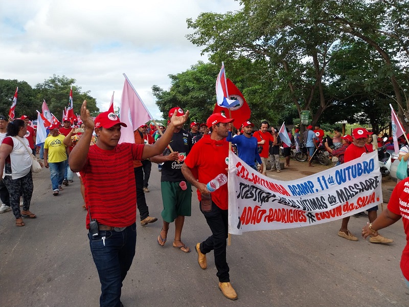 Grupo de manifestantes de vermelho marchando com bandeiras e um banner em protesto.