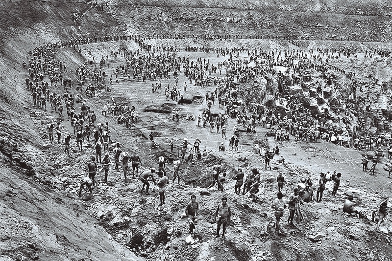 Homens trabalham em uma vasta mina a céu aberto, carregando pesados sacos, em fotografia em preto e branco.