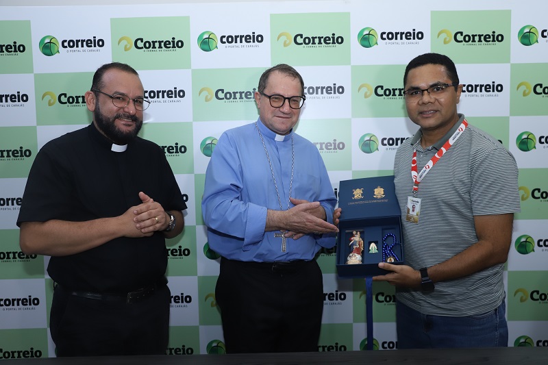 Três homens, incluindo um padre e um bispo, posam sorridentes com uma caixa de presente azul contendo uma estatueta religiosa e um terço azul.