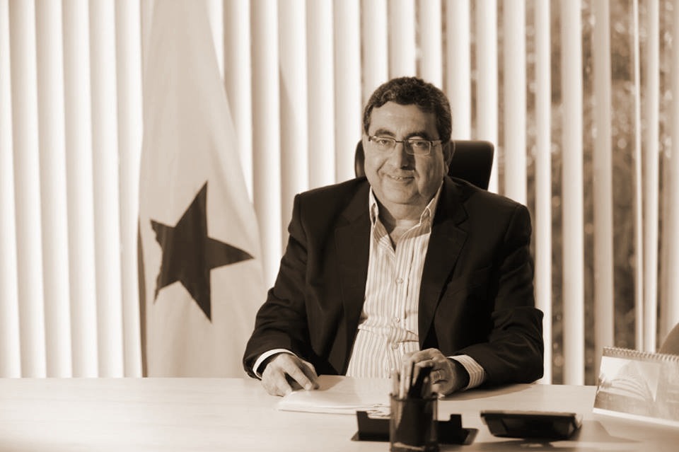 Executivo masculino de meia-idade sorrindo, sentado em um escritório com persianas.