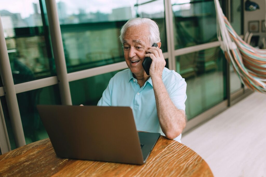 Homem idoso sorrindo e falando ao celular enquanto usa um laptop em uma mesa de madeira.