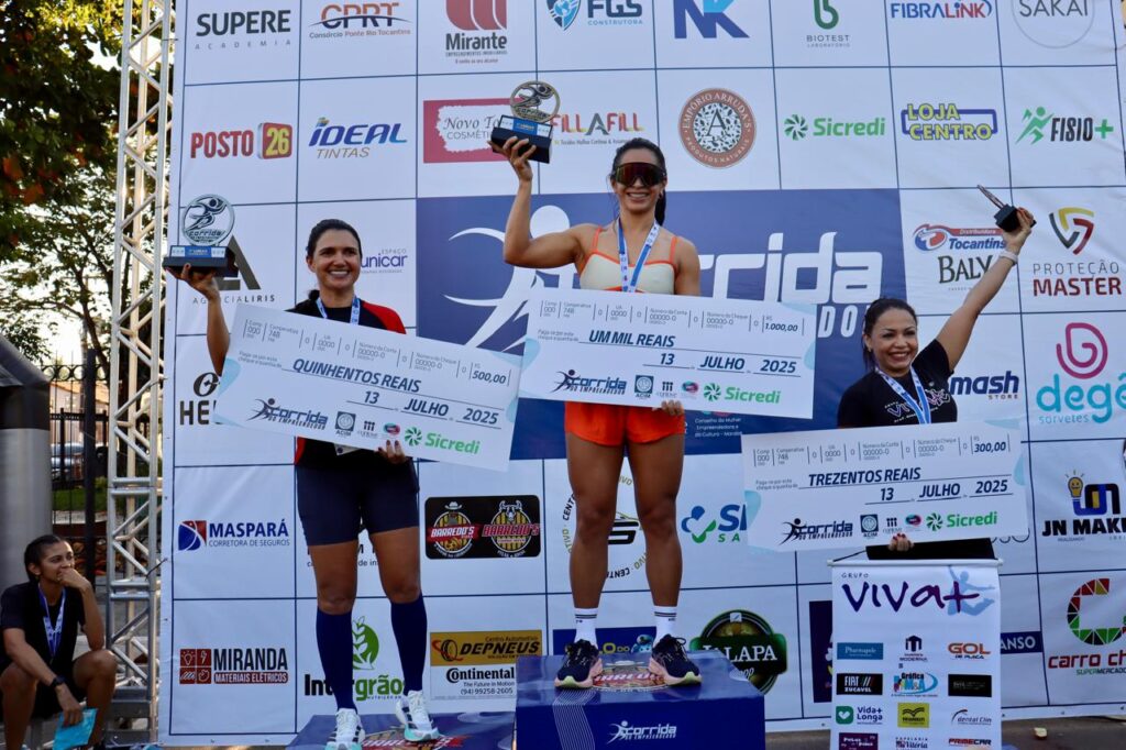Três mulheres atletas sorrindo no pódio de uma corrida de empreendedores, segurando troféus e cheques de premiação.