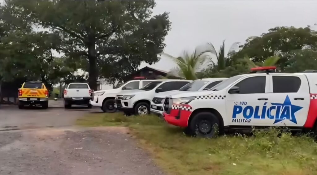 Fileira de caminhonetes brancas, incluindo uma viatura da Polícia Militar, estacionadas ao ar livre em um terreno com grama e terra.