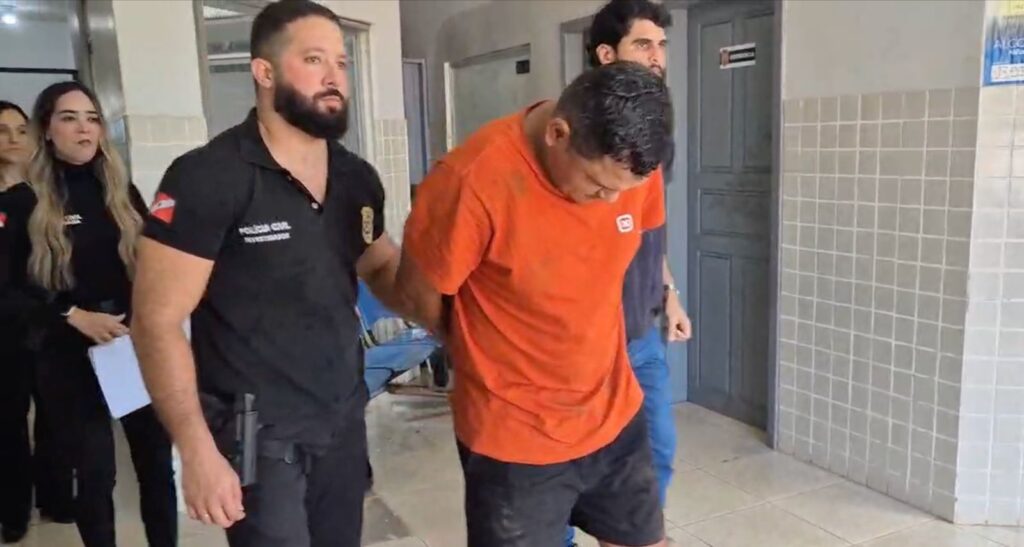 Homem de camiseta laranja algemado e escoltado por investigador da Polícia Civil.