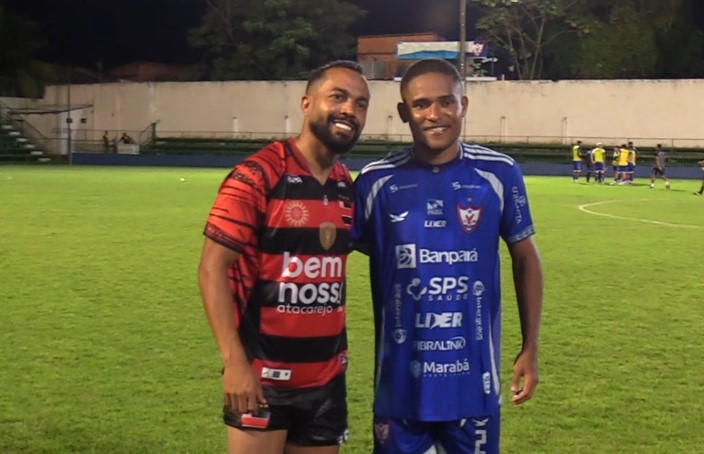 Dois jogadores de futebol sorrindo em campo de grama à noite.