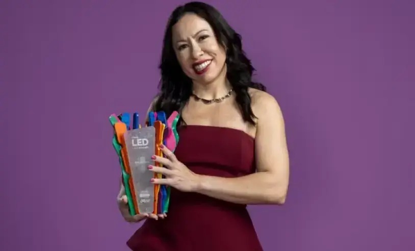 Mulher sorridente segurando um prêmio "LED" colorido contra um fundo roxo.