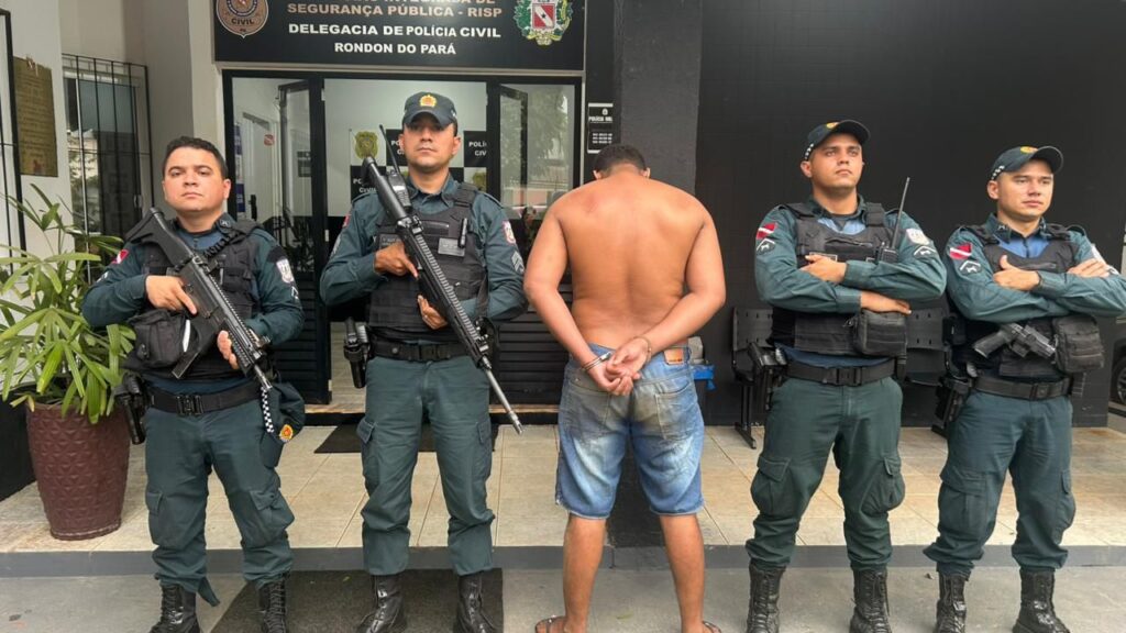 Homem algemado, sem camisa, entre dois policiais armados em frente à Delegacia de Polícia Civil de Rondon do Pará.
