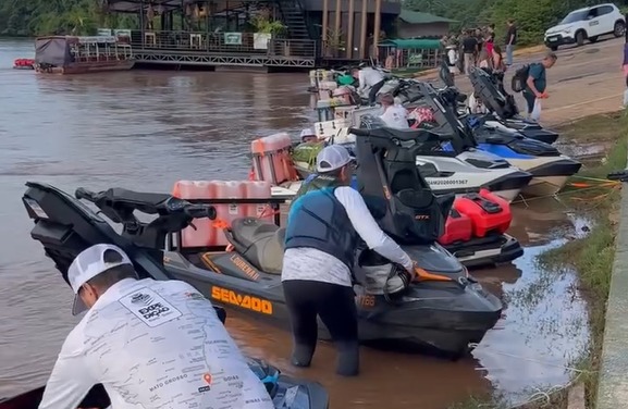 Jet skis estacionados na margem de um rio com pessoas se preparando e coletes salva-vidas visíveis.