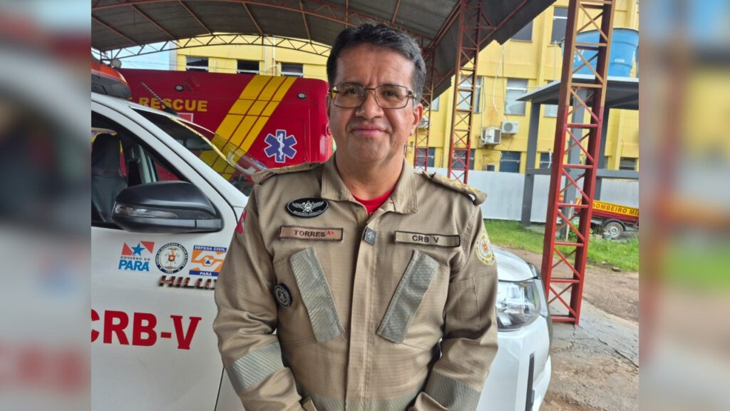 Homem de uniforme da Defesa Civil/Bombeiro em frente a um veículo de resgate.