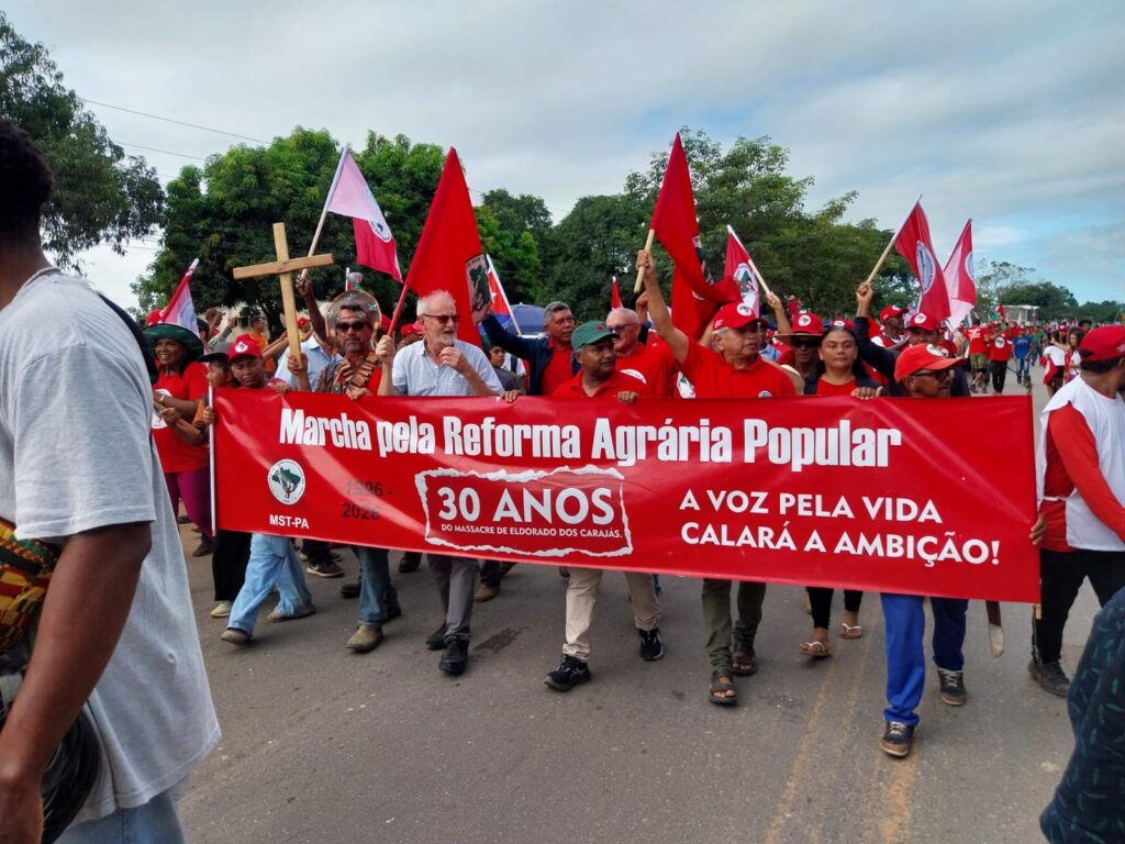 Grupo de manifestantes do MST em marcha com bandeiras e um banner vermelho "Marcha pela Reforma Agrária Popular".