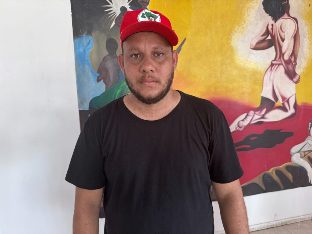 Homem com barba e boné vermelho com logo verde em frente a uma pintura colorida.