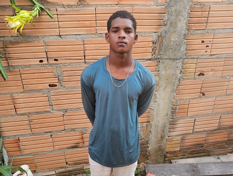 Jovem homem negro de camisa azul-petróleo e calça clara, em frente a uma parede de tijolos.