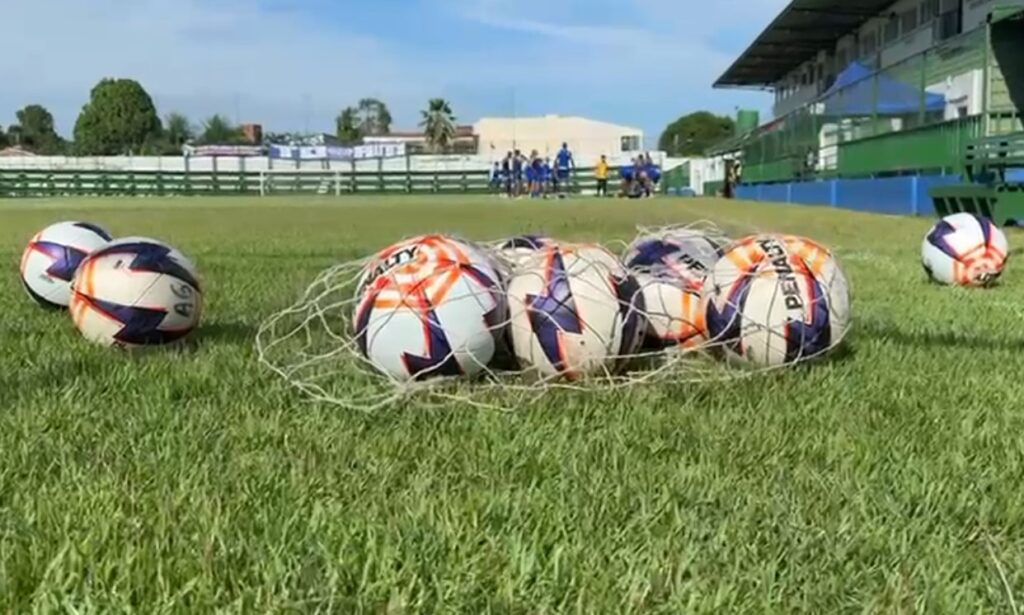 Bolas de futebol Penalty e Salty dentro de uma rede sobre a grama de um campo, com jogadores ao fundo.