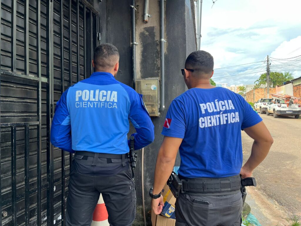 Dois policiais da Polícia Científica de costas, um de camisa de manga longa e outro de camiseta, em frente a uma parede escura com um medidor elétrico.