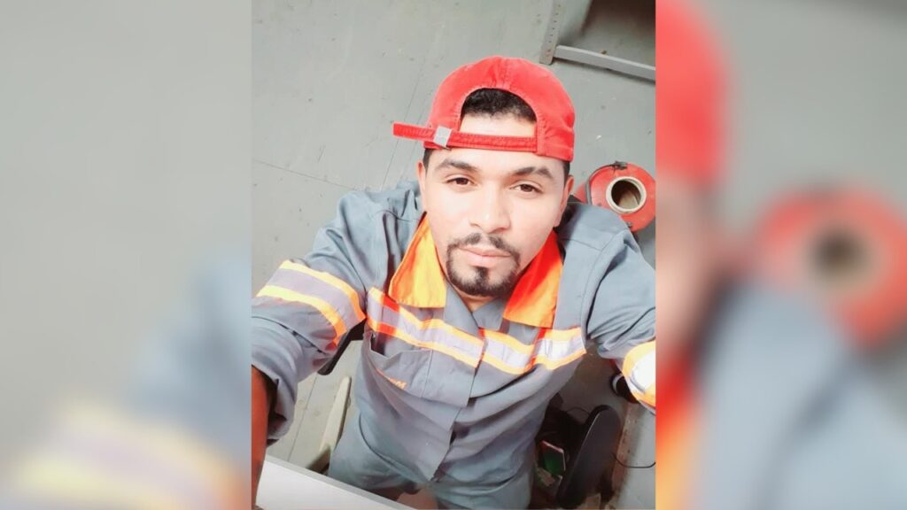Homem de uniforme de trabalho cinza com faixas refletivas e boné vermelho.