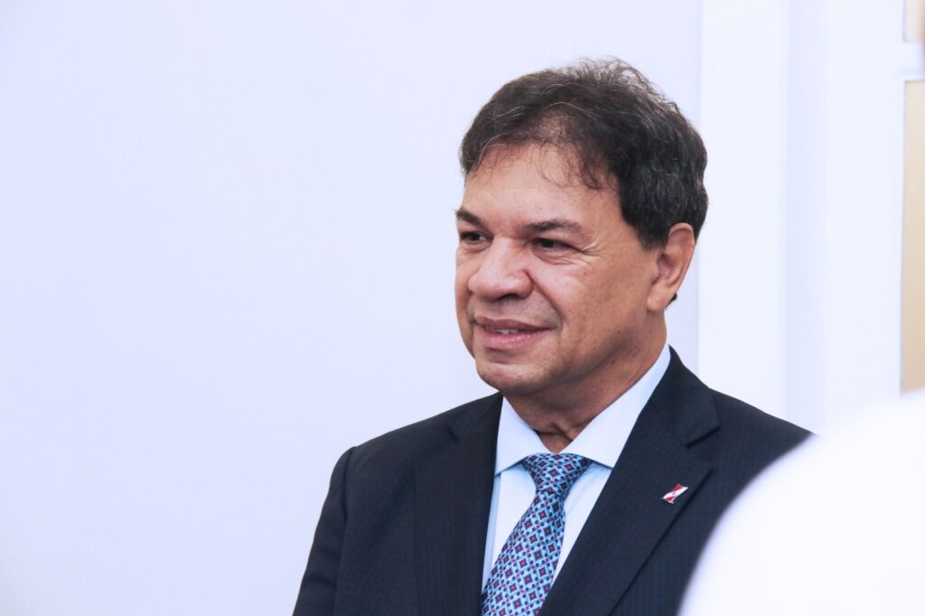 Homem de terno escuro e gravata azul sorrindo, com pino na lapela.