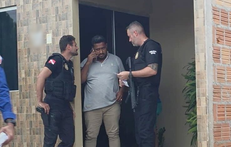 Policiais civis em operação abordam um homem falando ao celular na entrada de uma residência.