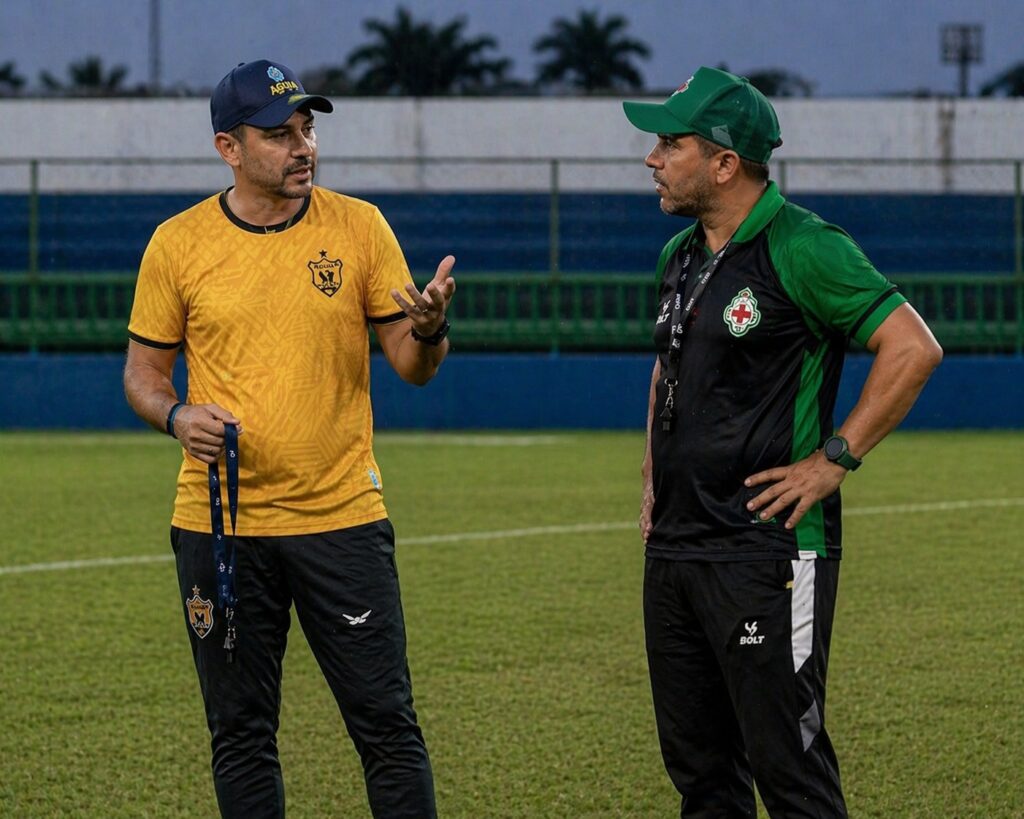 Dois homens em campo de futebol, um gesticulando enquanto o outro escuta.