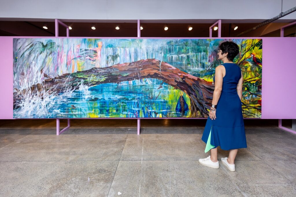 Mulher de vestido azul observando uma grande pintura abstrata colorida de paisagem natural com rio e galho.