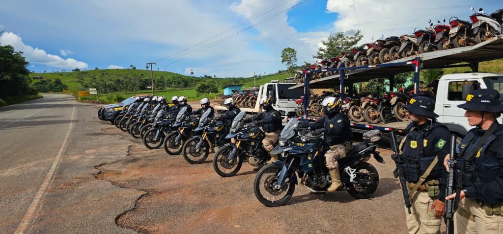 Fila de policiais da PRF em motocicletas de patrulha ao lado de um caminhão com motos apreendidas.