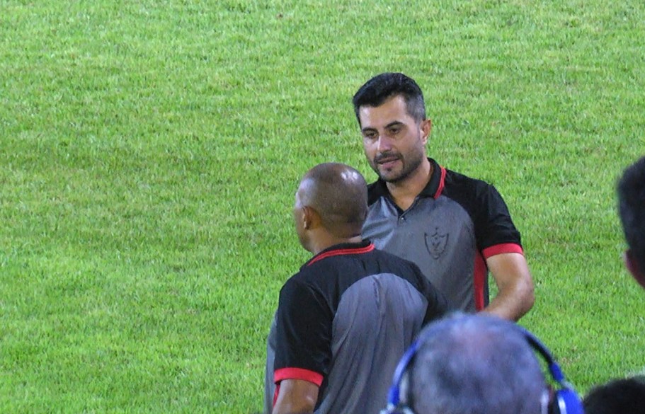 Homem de camisa polo com emblema em campo de futebol, conversando com outro de costas.