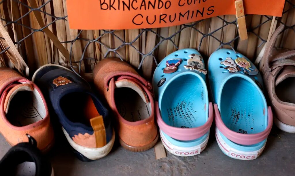 Sapatos infantis variados (um azul marinho, um laranja e dois Crocs azuis claros com enfeites) alinhados em frente a uma parede, sob um cartaz laranja escrito "BRINCANDO COM CURUMINS".