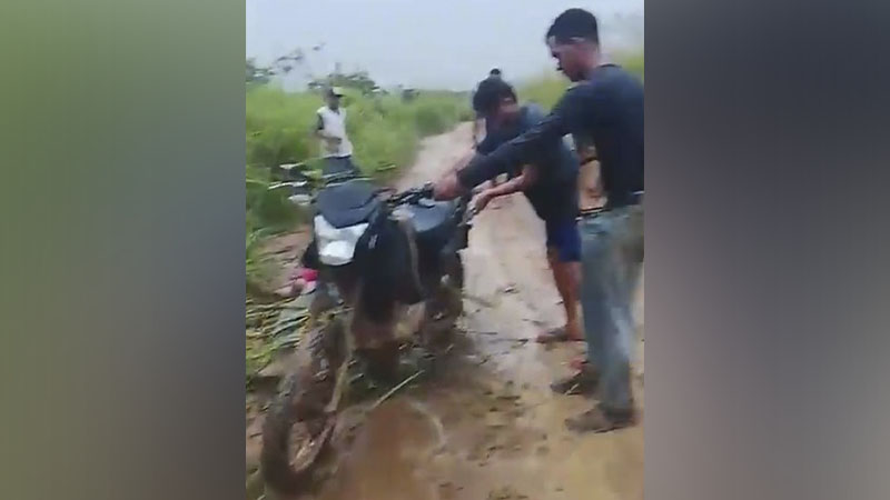 Dois homens empurrando uma moto preta atolada em uma estrada de terra lamacenta.