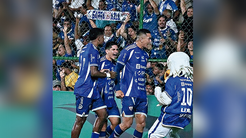 Jogadores de futebol em uniformes azuis e um mascote celebrando um gol em campo, com torcida animada ao fundo.