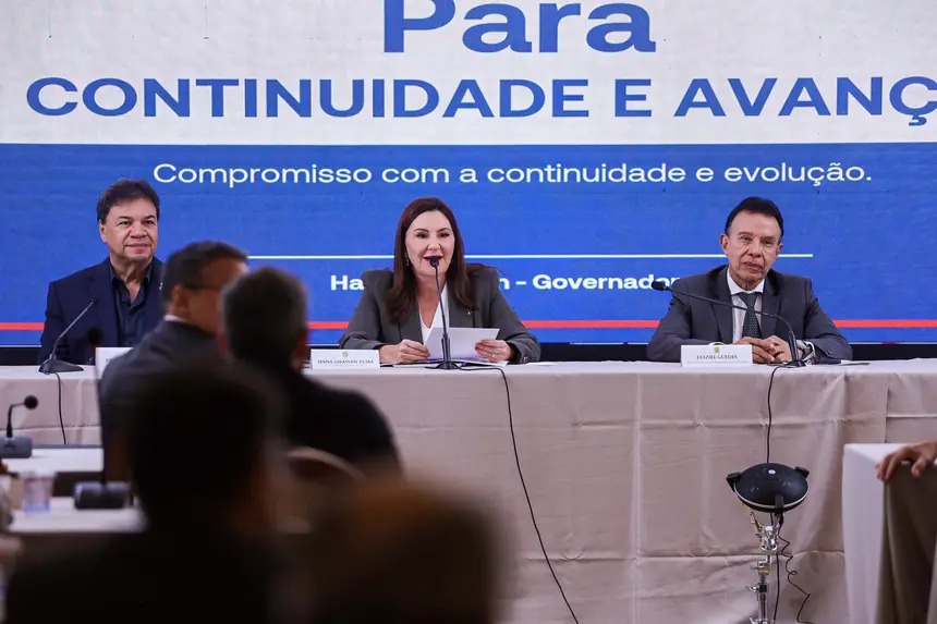 Mulher discursando ao microfone em evento político, com homem ao lado e banner azul de fundo.