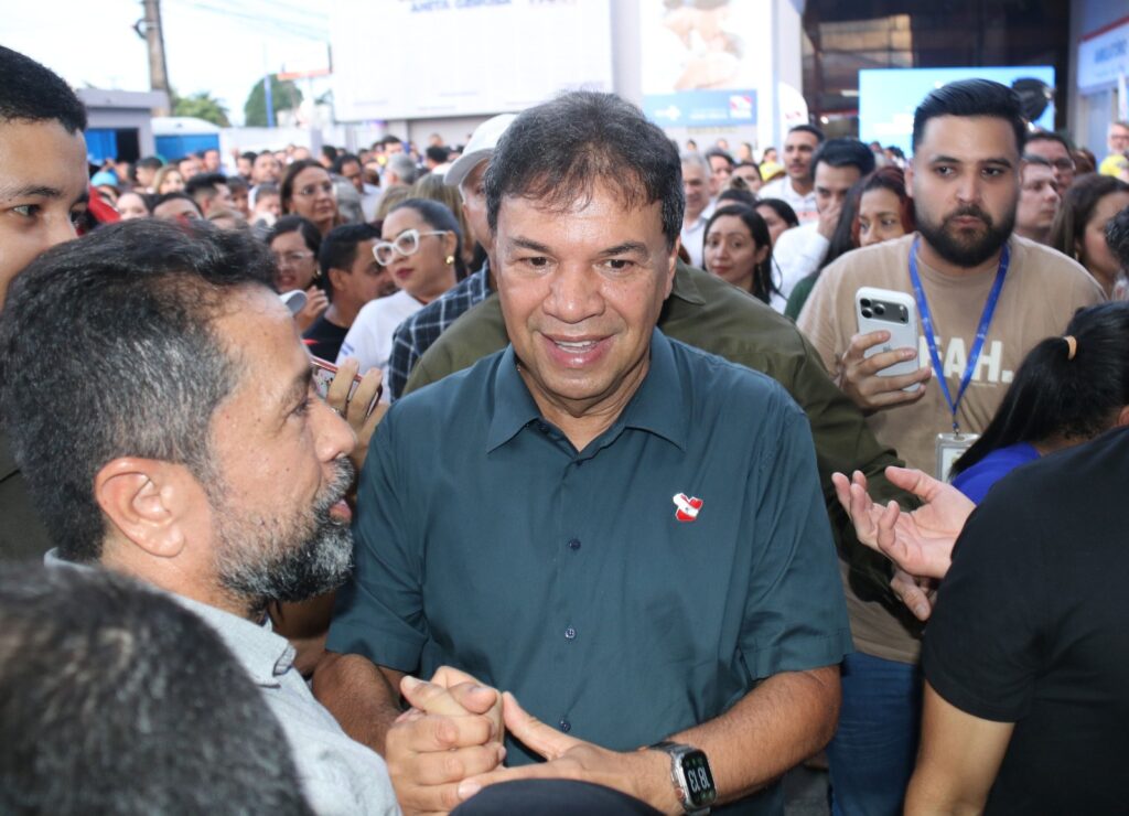 Homem sorridente de camisa verde-escura com broche em meio a uma multidão, interagindo com outra pessoa.