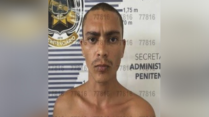 Homem de cabelo raspado e bigode fino em foto tipo "mugshot" contra parede com marcações de altura.
