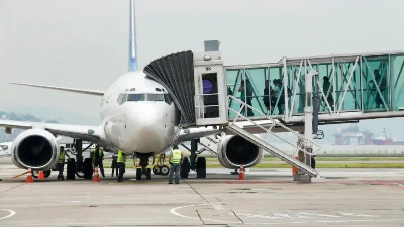 Avião branco estacionado no aeroporto com ponte de embarque e equipe de solo.