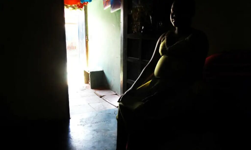 Mulher grávida em silhueta sentada em um quarto escuro com uma porta iluminada ao fundo.