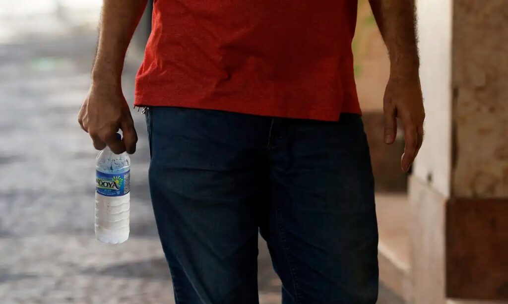 Pessoa de camiseta vermelha e calça jeans segurando uma garrafa de água.