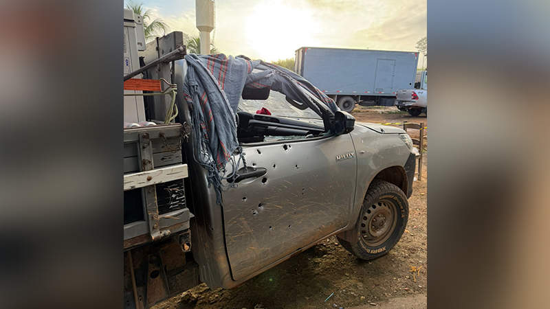 Caminhonete cinza com múltiplas marcas de tiros na porta e para-brisa estilhaçado.