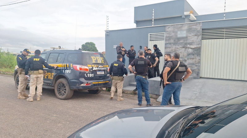 Oficiais da Polícia Rodoviária Federal (PRF) e outras forças de segurança reunidos em frente a uma residência, com um veículo da PRF estacionado.