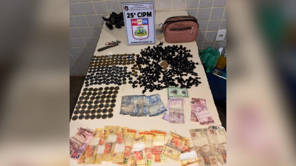 Mesa com grande quantidade de drogas (possíveis pedras de crack), dinheiro em notas e moedas, uma bolsa e um distintivo da 25ª CIPM.