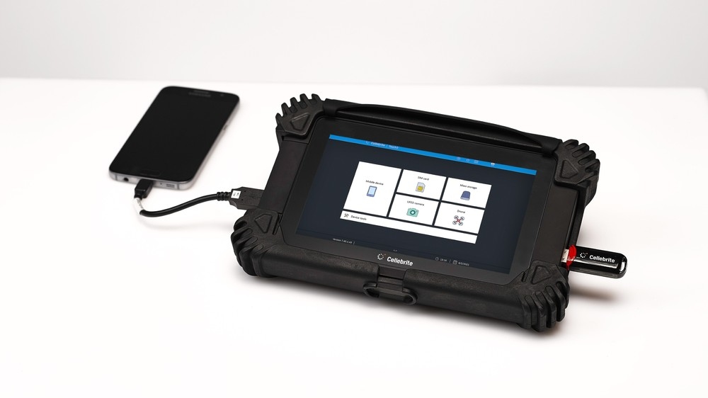 Tablet robusto Cellebrite conectado a um smartphone, exibindo software de extração de dados.