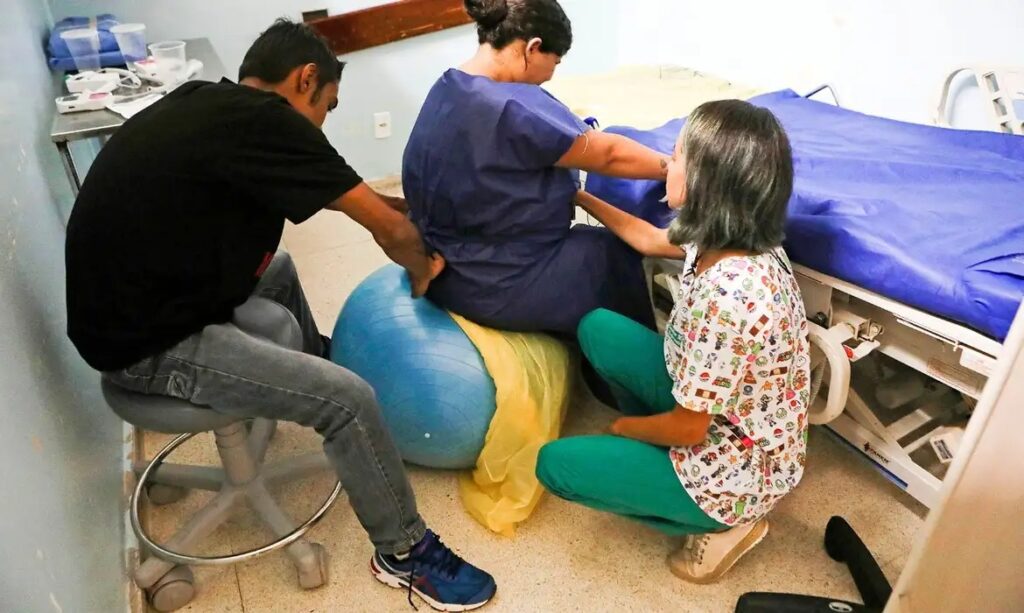 Mulher em trabalho de parto sentada em bola de fisioterapia, recebendo apoio do parceiro e de profissional de saúde.