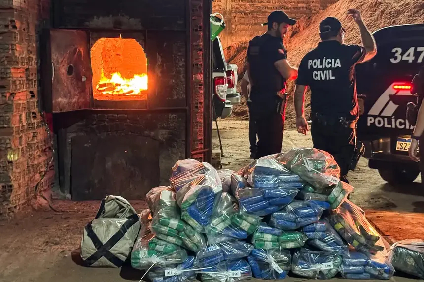 Pilha de pacotes de drogas aguardando incineração, com forno aceso e policiais civis ao fundo.