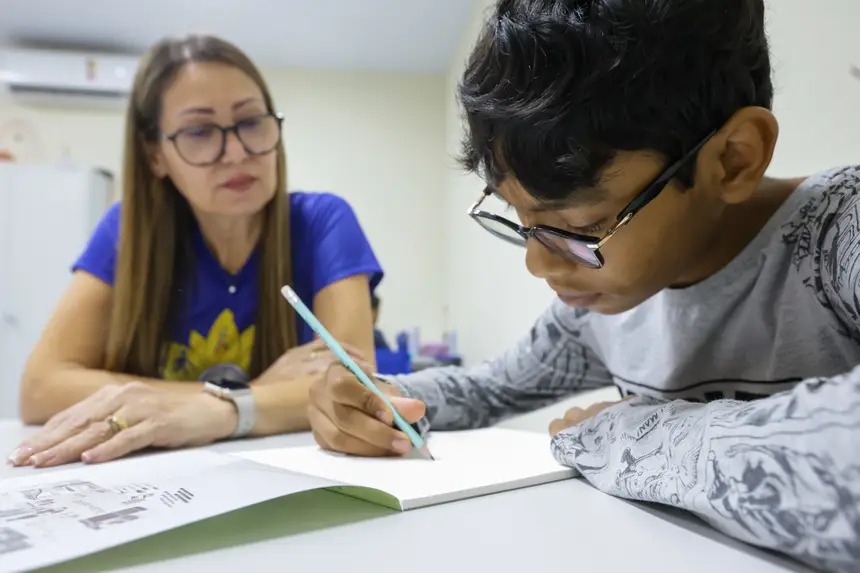 Menino escrevendo em caderno com supervisão de mulher.