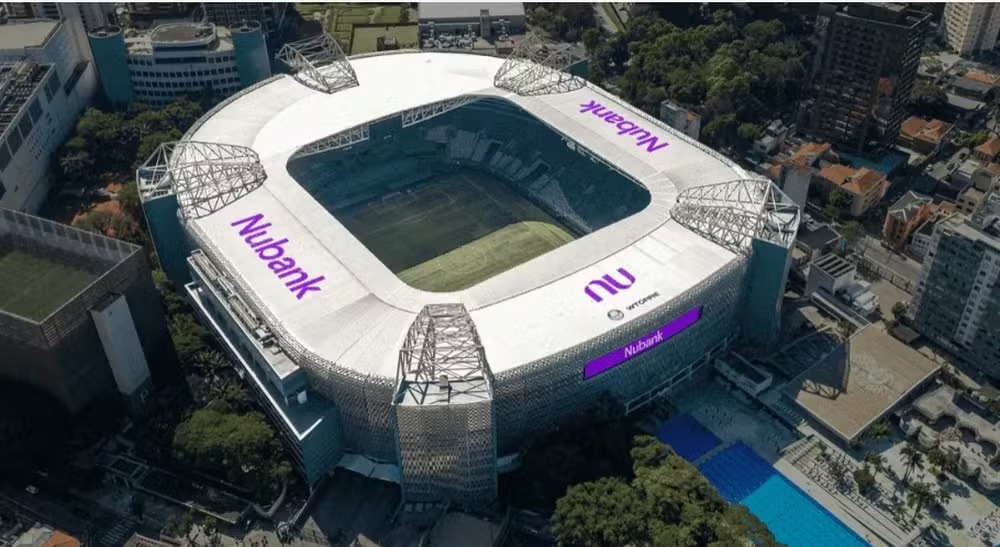 Estádio com a marca Nubank em roxo, visto de cima em área urbana.