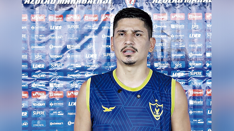 Homem em camisa de time de futebol azul e amarela posando em frente a um banner de patrocinadores.