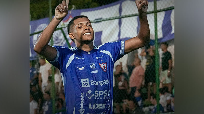 Jovem jogador de futebol em camisa azul comemorando com os braços erguidos e sorriso.
