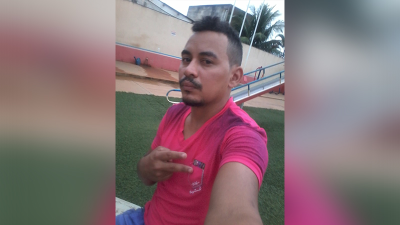 Homem de camiseta rosa fazendo sinal de paz em selfie ao ar livre.