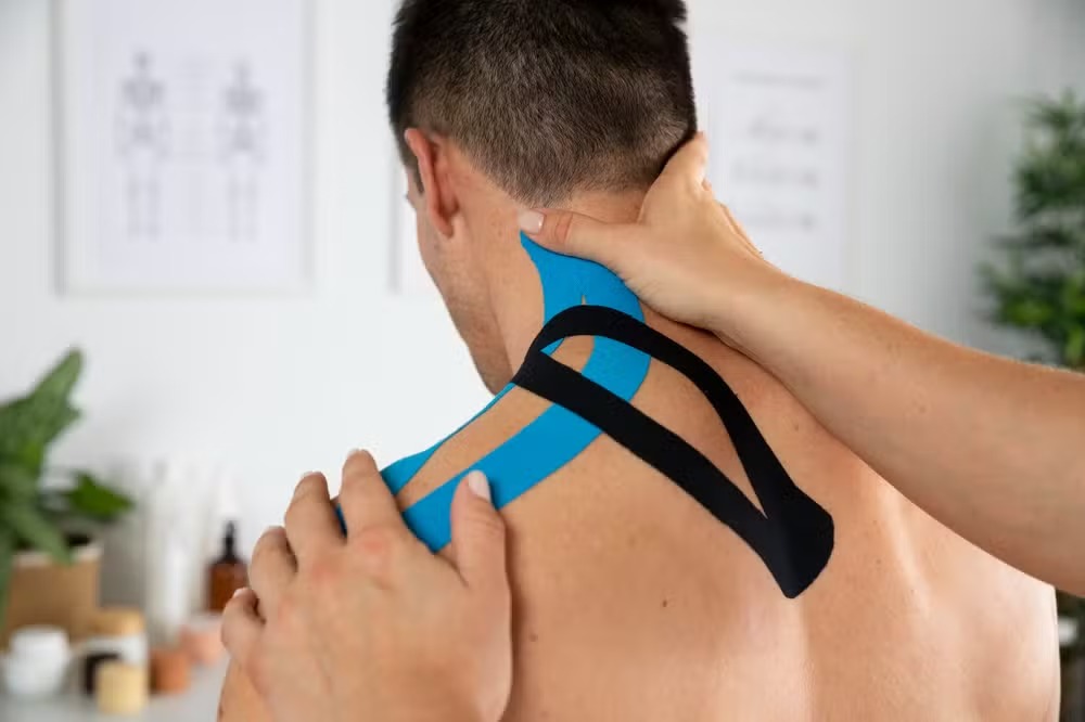 Fisioterapeuta aplicando bandagem elástica (kinesio tape) azul e preta no pescoço e ombro de um paciente.