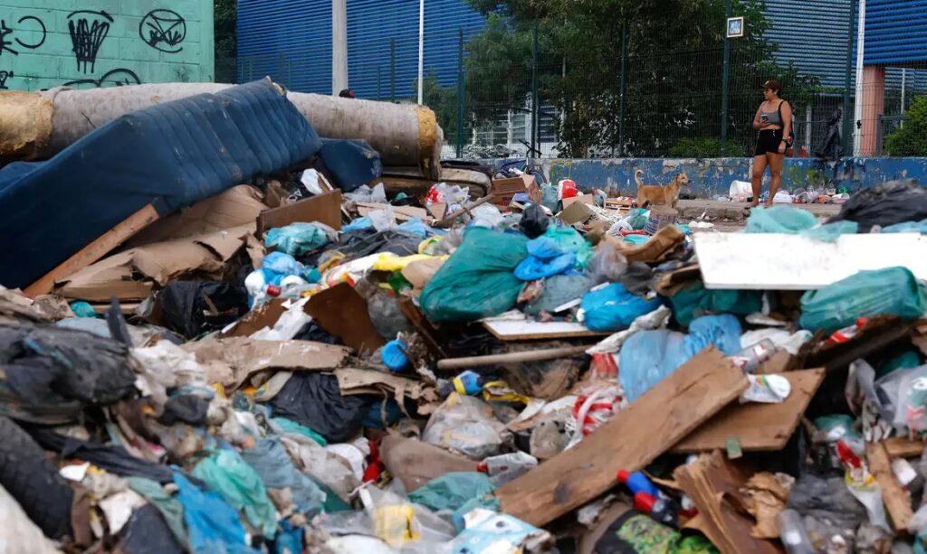 Grande monte de lixo e entulho em área urbana, com sofá velho e um cachorro ao fundo.