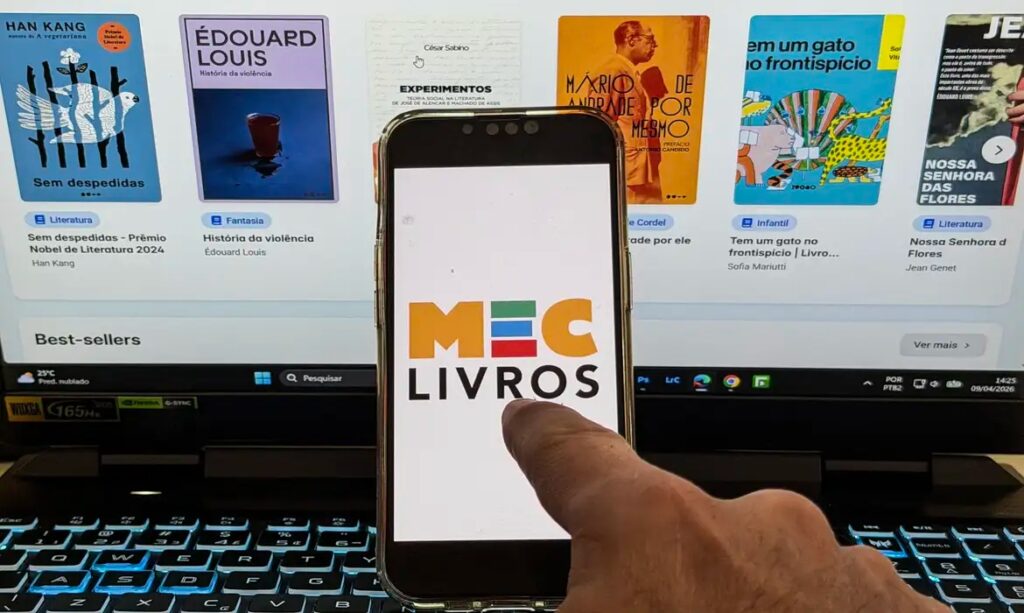 Mão segurando smartphone com app "MEC Livros" sobre tela de computador com livros digitais.