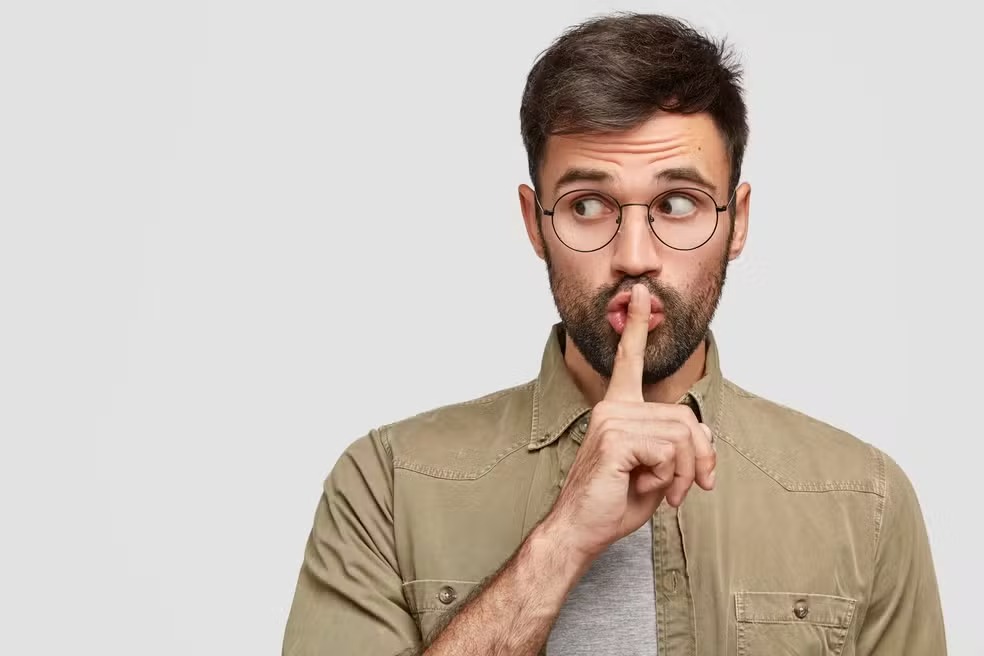 Homem com barba e óculos fazendo sinal de silêncio.